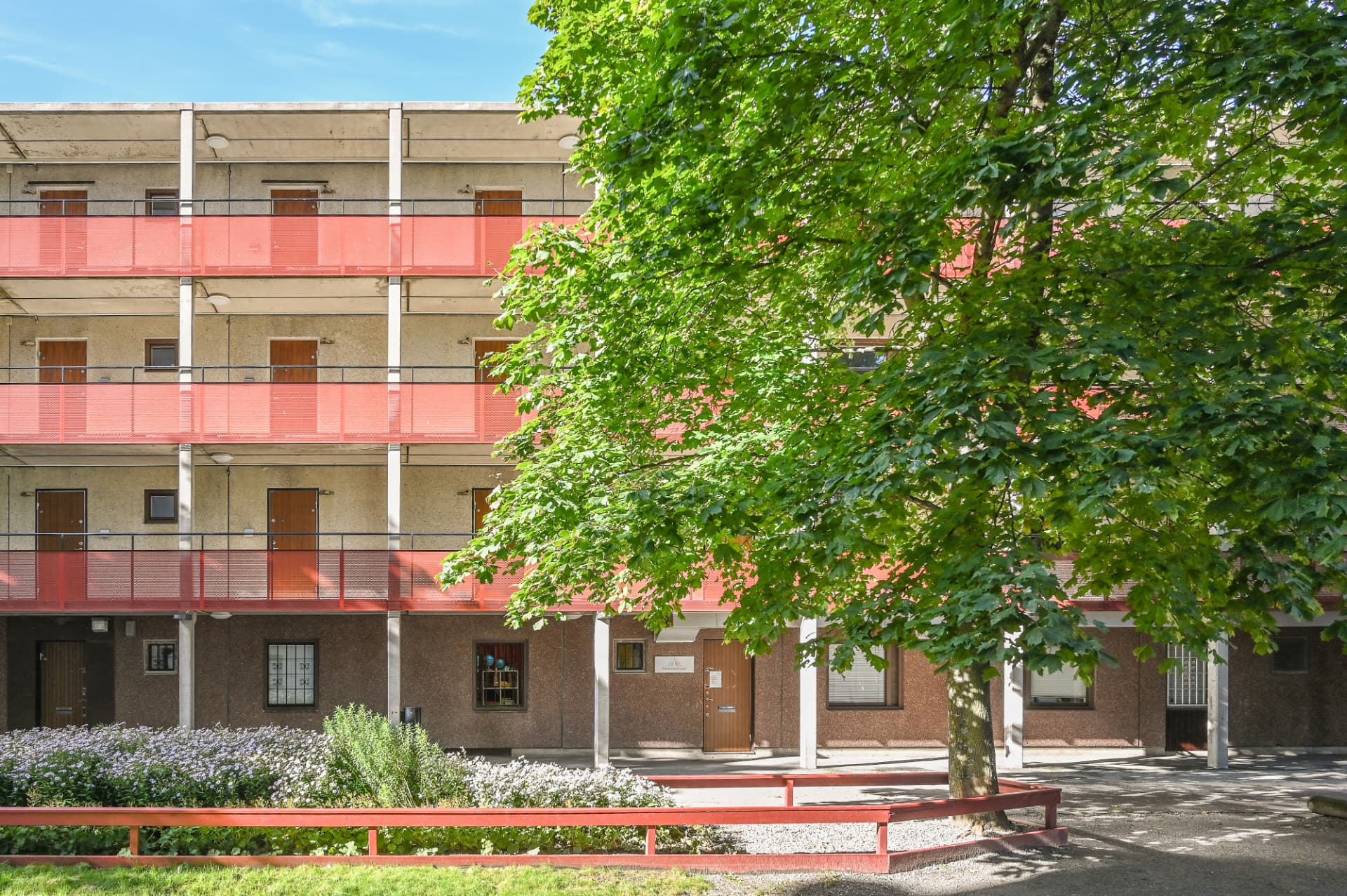 Förskolans entré i ett bostadshus med röda räcken och ett stort lövträd på framsidan
