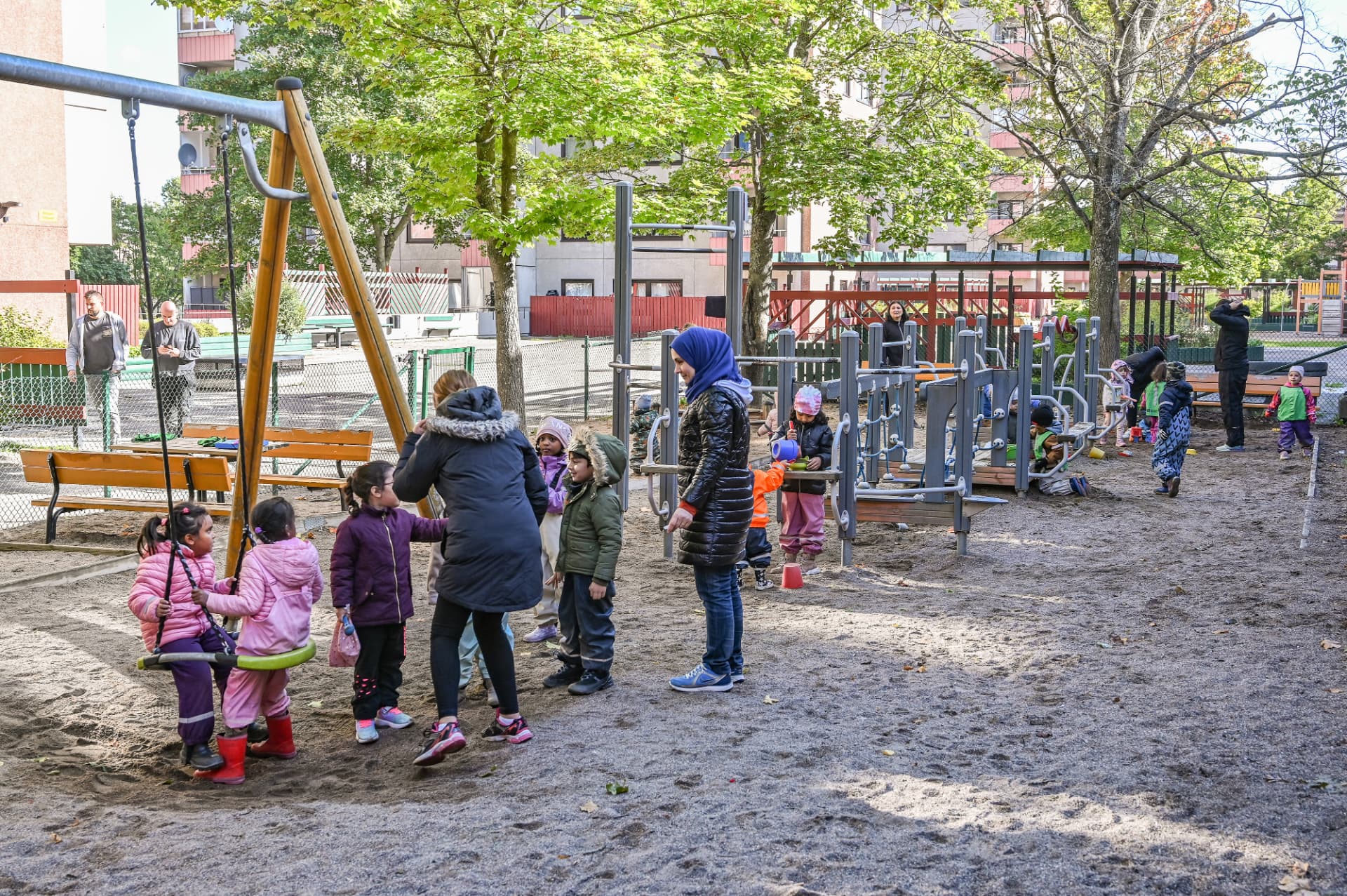 Barn och pedagoger på lekplatsen med gungor och klätterställning bland stora träd